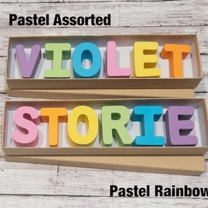 May include: Two boxes of wooden letters spelling out the words "VIOLET" and "STORIE". The letters are in pastel colors, with the top box labeled "Pastel Assorted" and the bottom box labeled "Pastel Rainbow".