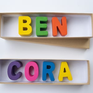 May include: Two boxes with the names "BEN" and "CORA" spelled out in colorful letters. The letters are made of wax and are arranged in the boxes.
