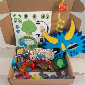 May include: A brown cardboard box filled with dinosaur-themed toys and activities. The box includes a coloring book, a felt dinosaur mask, a pop-it toy, and other items.