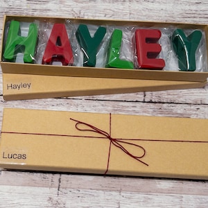 May include: Two brown cardboard boxes with red string tied around one of them. The top box is open and contains a set of colorful letters spelling out the name "Hayley". The bottom box has the name "Lucas" printed on it.