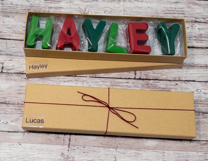 May include: Two brown cardboard boxes with red string tied around one of them. The top box is open and contains a set of colorful letters spelling out the name "Hayley". The bottom box has the name "Lucas" printed on it.