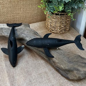 May include: Two black whale figurines with fins and tails are displayed on a piece of driftwood. The whales are positioned on a tan surface with a woven background. A potted plant in a wicker basket is in the background.