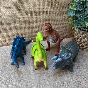 May include: Four toy dinosaurs in various colors. A blue Stegosaurus, a green and yellow Brachiosaurus, a brown Tyrannosaurus Rex, and a gray Triceratops. The toys are on a neutral-colored surface.