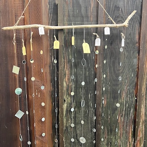 May include: A decorative mobile featuring a natural wood branch suspended by twine. Various shapes of reflective, silver-colored elements hang from the branch, creating a shimmering effect. The background is a weathered wooden fence.