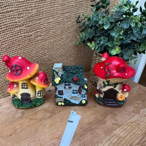 May include: Three whimsical mushroom houses with red caps and detailed facades. The houses are painted with vibrant colors and sit on a wooden surface. A small plant in a woven basket is in the background.