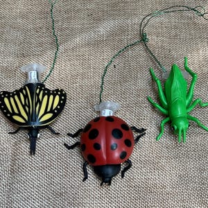 May include: Three insect-shaped ornaments featuring a yellow and black butterfly, a red and black ladybug, and a bright green grasshopper. Each ornament includes a clear plastic attachment point at the top connected to a thin green wire loop.