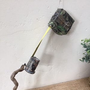 May include: A camouflage-patterned tape measure hanging from a ceiling, with a yellow tape extending down to a second camouflage-patterned tape measure. A brown, twisted branch is in the foreground.