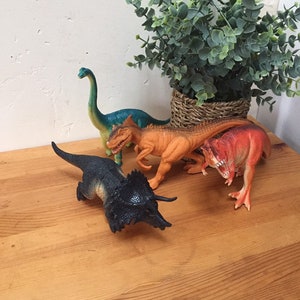 May include: Four plastic dinosaur toys on a wooden surface. The dinosaurs are a green and blue brachiosaurus, an orange and brown tyrannosaurus rex, a black and brown triceratops, and a red and orange velociraptor.