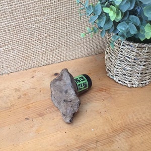 May include: A small, dark brown piece of driftwood with a green and black bottle stopper on top. The driftwood and bottle stopper are sitting on a light brown wooden surface.