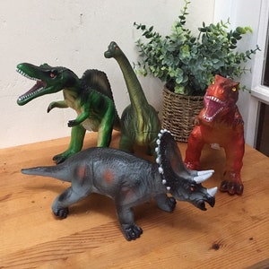 May include: Four plastic dinosaur toys, including a green Tyrannosaurus Rex, a green Brachiosaurus, a gray Triceratops, and an orange-red Tyrannosaurus Rex.
