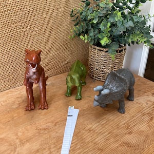 May include: Three toy dinosaurs: a brown T-Rex, a green dinosaur, and a gray Triceratops. The dinosaurs are on a wooden surface. A small piece of paper is in front of the dinosaurs. A woven basket with green plants is in the background.