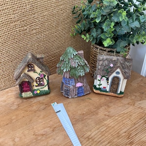 May include: Three miniature fairy houses with thatched roofs and detailed facades. One is beige with pink flowers, another is a tree-shaped house, and the third is a beige house with a brown door. The houses are on a wooden surface.