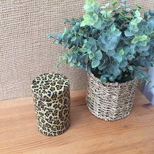 May include: A cylindrical container with a leopard print pattern sits on a wooden surface. The container is next to a small woven basket holding a green plant. The background is a textured, beige material.