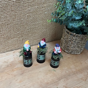 May include: Three miniature gnomes sitting on top of small green glass bottles. Each gnome is wearing a different colored hat: yellow, red, and purple. The gnomes are holding small gardening tools.
