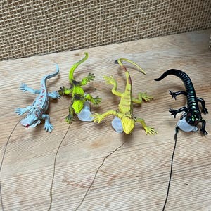 May include: Four toy lizards in various colors: light blue, green, yellow, and black. Each lizard has a different pattern and is attached to a thin wire. The lizards are lying on a wooden surface.