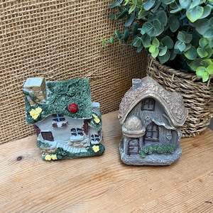 May include: Two miniature fairy houses. One has a green roof with a red accent and white walls. The other has a thatched roof and a stone facade. Both are detailed with windows and doors, perfect for a whimsical garden display.