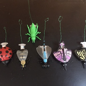 May include: A set of six colorful toy insects with strings attached. The insects include a ladybug, a bee, a dragonfly, a butterfly, a butterfly, and a grasshopper.