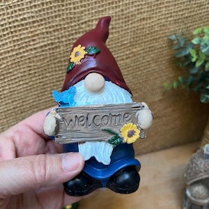 May include: A garden gnome figurine with a burgundy hat adorned with a yellow sunflower. The gnome holds a wooden sign that reads "welcome". A blue bird sits on the sign. The gnome wears a blue outfit and black shoes.