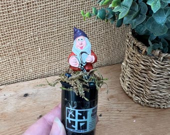 May include: A whimsical garden gnome figurine with a purple hat and red coat, holding a pair of silver spectacles, atop a black container with text and a graphic. The container is decorated with moss.