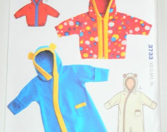 Op de afbeelding: Kwik Sew babykledingpatroon, maat XS-XL. Het patroon toont illustraties van babyjassen en een slaapzak. De jassen zijn rood met blauwe afwerking, en een rode jas met kleurrijke stippen. De slaapzak is blauw met gele afwerking.
