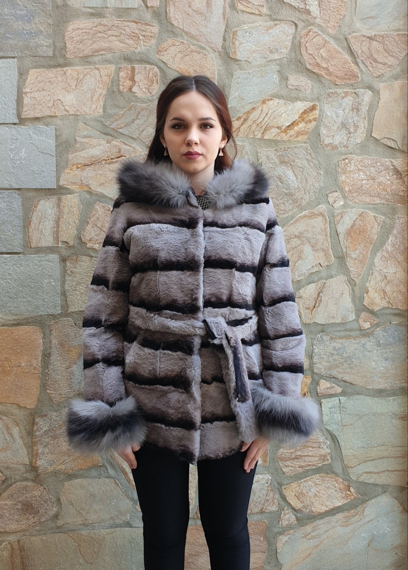 Puede incluir: Un abrigo de piel de color gris y negro a rayas con capucha y pu&ntilde;os forrados de piel. El abrigo tiene un cintur&oacute;n negro y est&aacute; ce&ntilde;ido a la cintura.