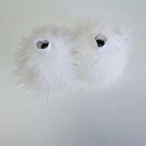 Pode incluir: Dois chinelos brancos fofos com uma textura felpuda. Cada chinelo tem uma faixa elástica branca ao redor da abertura e um interior escuro. Os chinelos estão em uma superfície branca.