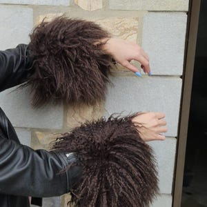 Pode incluir: Um casaco de couro preto com punhos longos de pelo sintético encaracolado castanho-escuro. As mangas do casaco são feitas de material preto liso com um detalhe de fecho de metal prateado visível perto do pulso, acima do acabamento em pelo.