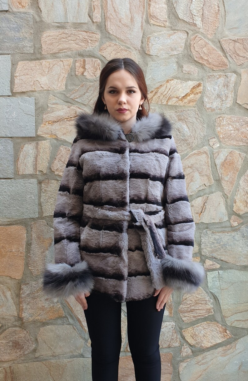 Puede incluir: Un abrigo de piel de color gris y negro a rayas con capucha y cintur&oacute;n. El abrigo tiene un ribete de piel negra alrededor de la capucha y los pu&ntilde;os.
