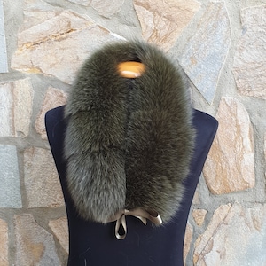 Pode incluir: Gola de pele verde-oliva com laço de fita de cetim. Apresentada num manequim preto, com um fundo de parede de pedra. A pele tem uma textura macia e fofa, com um aspeto natural e luxuoso.