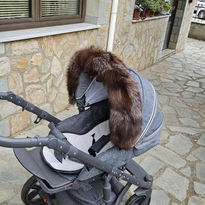 Puede incluir: Un cochecito gris con un marco negro y un asiento de tela blanco y gris. Una capucha de piel marrón y plateada está unida al cochecito.