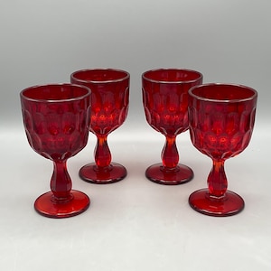 May include: Four ruby red wine glasses with a textured pattern. Each glass has a rounded bowl, a stem, and a circular base. The glasses are arranged on a white surface.