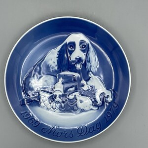 May include: A blue and white decorative plate featuring a dog with puppies and a basket. The plate has the words "1969 Mors Dag 1979" in a cursive font around the edge. The plate has a white rim.