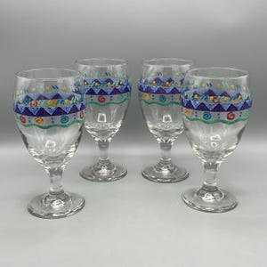 May include: A set of four clear glass goblets featuring a hand-painted decorative band around the upper rim. The design includes a row of purple triangles, a green wavy line, and small colorful swirls in red, yellow, and blue accents.