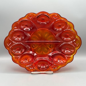 May include: An orange-red glass serving dish with a starburst design. The oval-shaped dish has a central section and multiple oval compartments around the edge. The dish is supported by a white stand.