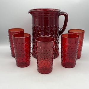May include: A deep red glass pitcher and four matching tall tumblers, all featuring a raised hobnail texture across their exterior surfaces. The pitcher includes a wide pouring spout and a sturdy side handle, while the glasses are cylindrical.