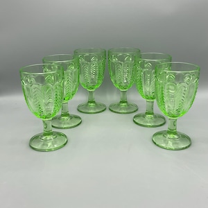 May include: A set of six green-tinted glass goblets. Each goblet features a detailed, repeating leaf pattern on the bowl and a rounded base. The glasses are arranged in a semi-circle, showcasing their design and color.