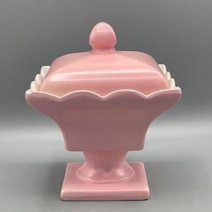 May include: A square pink ceramic candy dish with a matching lid featuring a pointed finial. The container has a scalloped upper rim with white detailing and rests on a pedestal base that transitions into a square footed bottom section.