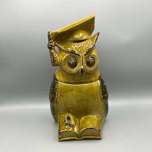 May include: A ceramic owl-shaped cookie jar in shades of yellow and black. The owl wears a graduation cap and holds a tassel. The owl is perched on an open book. The jar has a lid and is likely used for storing cookies or other treats.