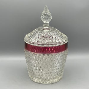 May include: A clear glass jar with a diamond pattern and a ruby red band around the middle. The jar has a matching lid with a decorative finial. The jar is likely used for storage or display.