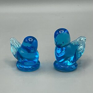 May include: Two small, blue glass bird figurines. The birds have rounded bodies, small beaks, and clear, wing-shaped details. The figurines are a vibrant shade of blue and sit on a light gray surface.