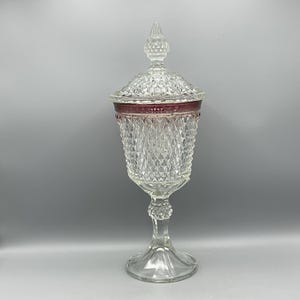 May include: A tall, clear glass candy dish with a lid. The dish has a diamond pattern and a red band around the top. The lid is topped with a decorative finial. The dish stands on a pedestal base.