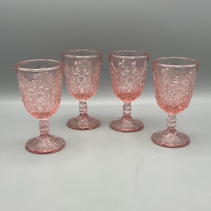 May include: Four pink-tinted stemmed glasses. Each glass features a detailed, embossed pattern on the bowl and base. The glasses are arranged in a row, showcasing their uniform design and color. Ideal for serving beverages.