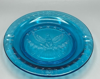 May include: A turquoise glass ashtray with a detailed embossed design. The center features an eagle with outstretched wings, surrounded by stars. The rim has a floral and leaf pattern. Two cigarette rests are visible.