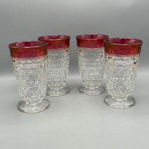 May include: A set of four clear glass tumblers with a textured hobnail pattern and a deep red band around the top rim. Each glass features a sturdy circular base and a slightly flared shape, showcasing a consistent design across all four pieces.