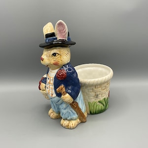 May include: A ceramic bunny planter with a blue jacket, a brown hat, and a brown stick in its hand. The planter has a woven basket design and is decorated with green leaves and red flowers.