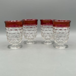 May include: A set of four clear glass tumblers with a textured diamond pattern on the body and a deep red band around the rim. Each tumbler features a short, sturdy pedestal base, providing a stable foundation for the faceted, transparent vessel.