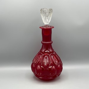 May include: A ruby red glass decanter with a clear, ornate stopper. The decanter has a bulbous body with a textured pattern and a long neck. The stopper is detailed with vertical lines and a floral design.