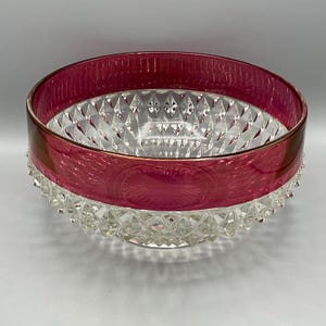 May include: A clear glass bowl with a ruby red band around the top. The bowl has a diamond-cut pattern on the inside and a textured, spiky base. The rim of the bowl has a gold-colored trim.