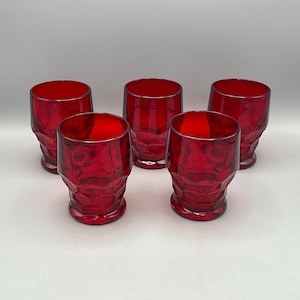 May include: Five red drinking glasses with a textured, faceted design. The glasses have a deep red color and a silver rim. The glasses are arranged in a group, showcasing their unique shape and color.