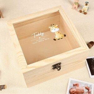 May include: A light-colored wooden keepsake box with a clear glass top. The box features a metal clasp and a giraffe sticker. Text on the glass reads "Teddy" with a date, time, weight, and height. A baby photo and ultrasound image are nearby.